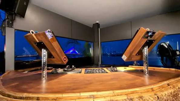 A curved wooden rooftop booth with two angled timber DJ stands and a turntable between them, city lights and water visible through the windows behind.