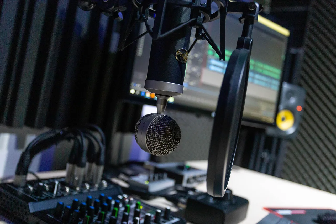 A condenser microphone on a boom arm with a pop filter in front of it, a mixing desk and patch cables below, a monitor and acoustic foam panels behind, all lit low and blue.