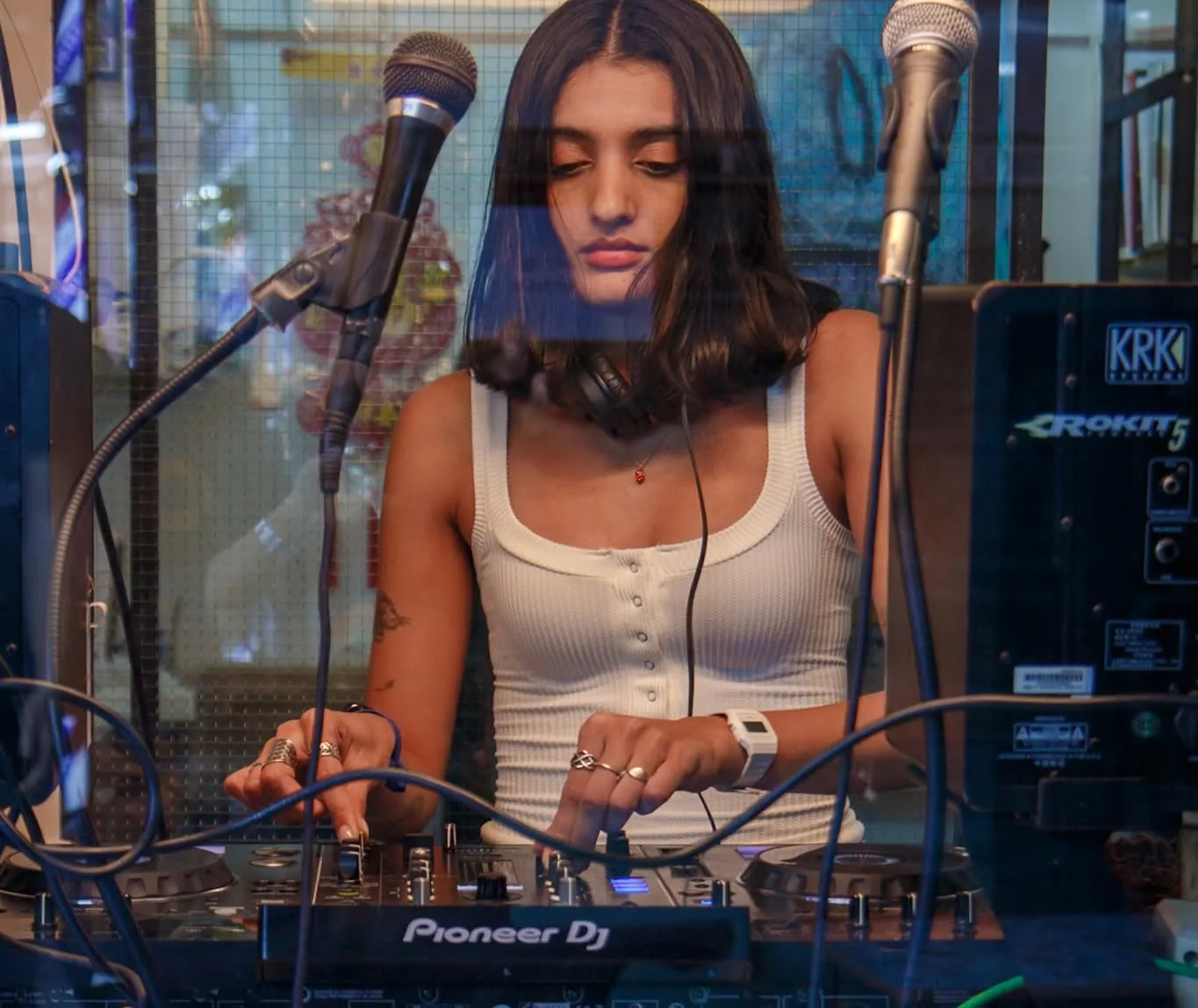 A DJ mixing on a Pioneer setup, seen from outside through the studio glass, two microphones overhead and street reflections across the window.
