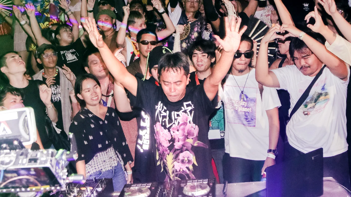 A crowd packed tight around a DJ booth with hands and phones in the air, the DJ mid-set with both arms raised.