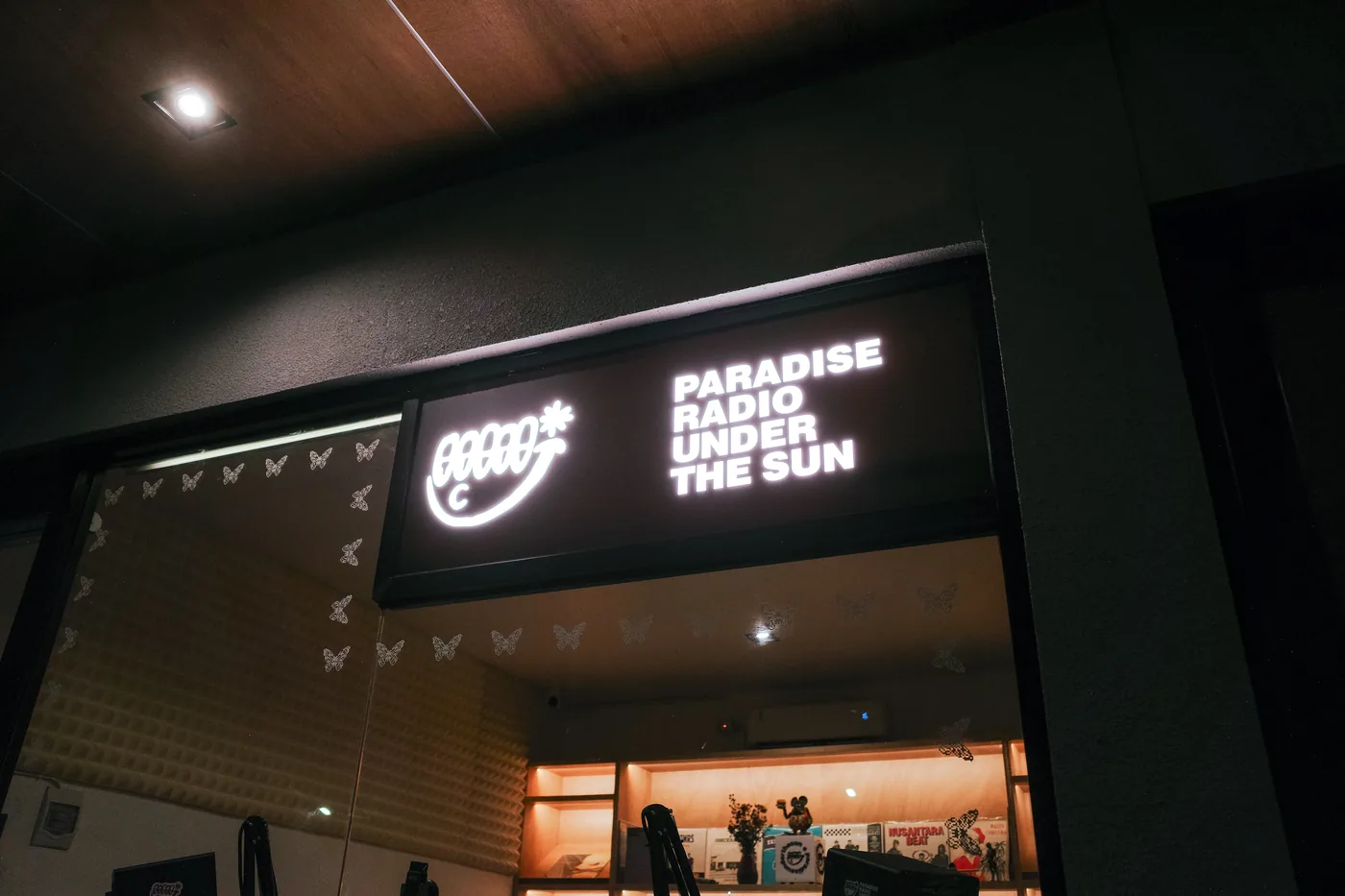 The Paradise Under the Sun shopfront after dark, an illuminated sign reading PARADISE RADIO UNDER THE SUN above a glass studio front, warm light and shelving visible inside.
