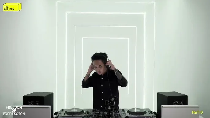 A DJ in a white room, hands to their headphones behind a pair of decks, framed by concentric strips of white light on the wall behind.